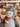 A plush donkey toy with gray and white fur, wearing a red and white Christmas tree patterned scarf, photographed in a shop setting with shelves of other toys blurred in the background.