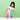 A young girl with dark hair wearing a white apron over a light purple t-shirt is smiling and interacting with a colorful plastic marble run toy set on a white table against a solid light green background.