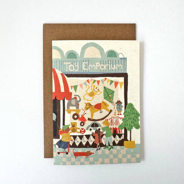 A greeting card with a kraft brown envelope, featuring a whimsical illustration of a toy store called 'Toy Emporium' with playful characters and colorful items, set against a plain white background.