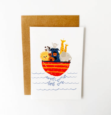 A white greeting card with a brown kraft envelope behind it, featuring a colorful illustration of safari animals in a boat with the words 'welcome little one' in blue script below.