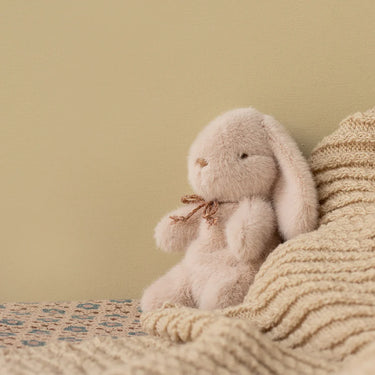 A soft plush bunny toy in a pale blush pink color sits on a textured knit blanket against a solid beige background.