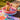 A young child stands in an inflatable pool, holding a toy water sprayer that is spraying water. The pool is set on a colorful play mat on a wooden deck next to a swimming pool.