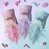 Three stand-up pouches of Magic Powder Cosmetics Wonder Poudré Mix are arranged on a pastel surface, with pink, purple and blue powder spilled below each pouch. Decorative squiggle shapes are placed around the scene.