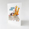 A white greeting card standing upright on a light gray surface, featuring a colorful cartoon illustration of safari animals including giraffes, an elephant, a zebra, a monkey, and a pink pig, with a blue banner reading 'Happy Birthday'.
