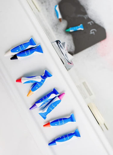 A set of bath crayons shaped like fish, displayed on a white surface with a blurred sink and soap dish in the background.