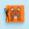 An orange felt activity book featuring a brown bear face with its eyes covered by its paws, tied with a black ribbon on a light blue background.
