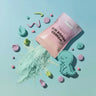 A pink stand-up pouch labeled 'WONDER DOUGH MIX' lies on a light blue surface with pale mint powder spilled in front. The scene is styled with pastel dough-like shapes and a mint-green dinosaur cookie cutout.