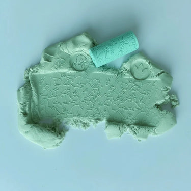 Blue play dough with embossed dinosaur motifs, shown with a matching mint green cylindrical chisel tool on a light blue background.