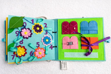 A colorful children's felt activity book or sensory pocket album shown open on a white textured background. The left page has numbered flower cutouts and ribbon loops, while the right page has fabric pockets in pink, blue, purple, and orange with ribbons.