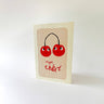 A greeting card with a cream border and a central beige panel featuring two red cherries with black stems and the French phrase 'mon chéri' in red script, photographed on a clean white background.