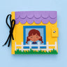 A handmade felt jbusy book featuring a house design with a purple scalloped roof, yellow body, white picket fence, green grass, and a cartoon girl in a blue window frame. A black ribbon is tied to the left side, and the item is photographed on a light blue background.