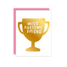 A square greeting card with a white front featuring a large gold trophy graphic and the words “MOST AWESOME FRIEND” in white. A pink card back or second card is visible behind it on a clean white background.