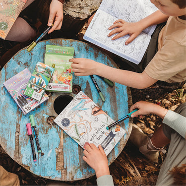 Kids sitting around a round wooden table with an activity boo, textas, pencils on a bark and leaf strewn forest floor