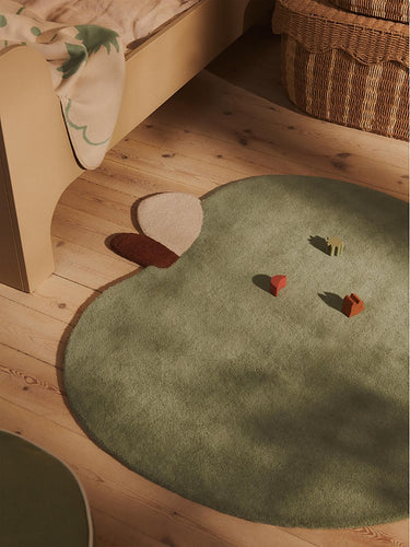An Ferm Living organic-shaped sage green children's rug with animal-like ears and small acorn details, styled on a light wood floor in a cozy room next to a bed.