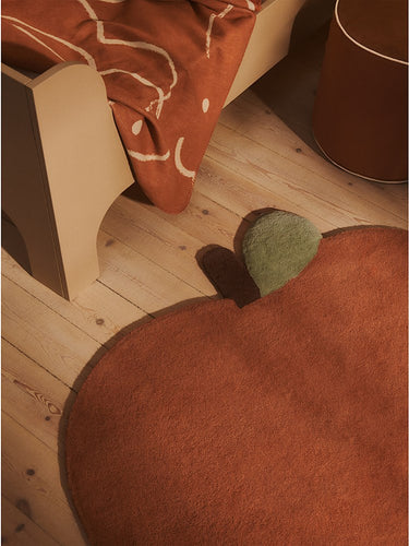 A close-up of a Ferm Living rust-orange  rug on a light wood floor, with part of a wooden bed frame and an orange patterned blanket visible in the room.