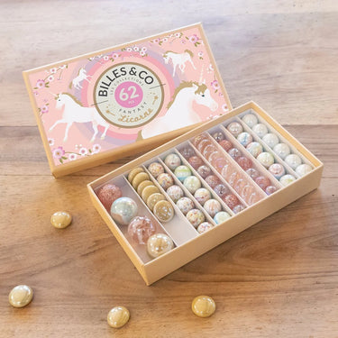 An open wooden box of Billes & Co. Unicorn themed marbles on a wooden tabletop, with the pink unicorn-patterned lid placed beside it and several loose marbles scattered around.