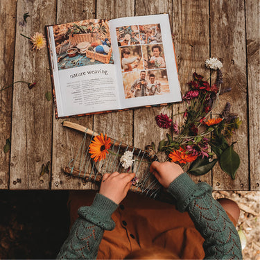 A child making a craft project on a wooden table outside, with a book open in front of her