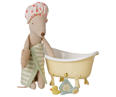 A plush toy mouse dressed as a bath bunny, wrapped in a green and white striped towel and wearing a pink polka-dot shower cap, sitting next to a pale yellow clawfoot bathtub. A small blue and gold bath bottle, a yellow rubber duck, and a brush are placed in front on a white background.