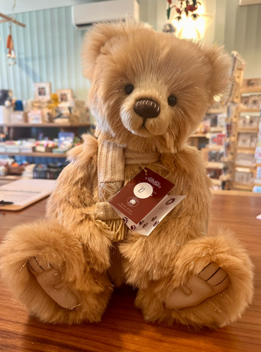 A plush teddy bear with light brown fur with glittery silver accents sits on a wooden table in a gift shop. The bear is wearing a cream-colored knit sweater and has a hangtag attached to its neck. He is labelled Bear B.