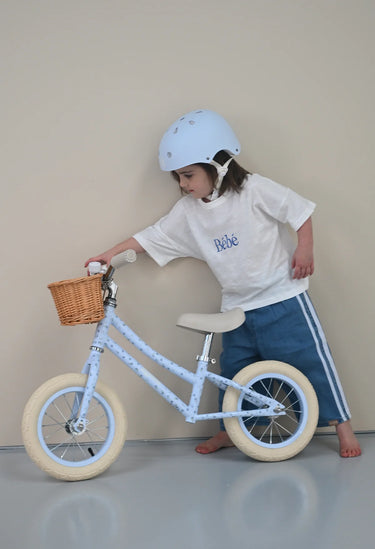 A child wearing a white helmet, white t-shirt with 'Biche' printed on the chest, and blue shorts stands next to a light blue balance bike with cream tires and a wicker basket, set against a plain beige wall.