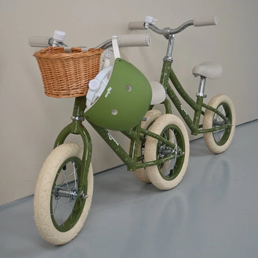 An olive green children's balance bike with cream tires, a wicker basket, and a matching green helmet, photographed indoors against a plain beige wall and gray floor.