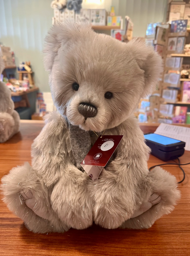 A plush teddy bear with light gray and sparkling fur sits on a wooden table in a gift shop. The bear has a dark gray winter scarf and a red hangtag attached to its neck. He is labelled Bear B.