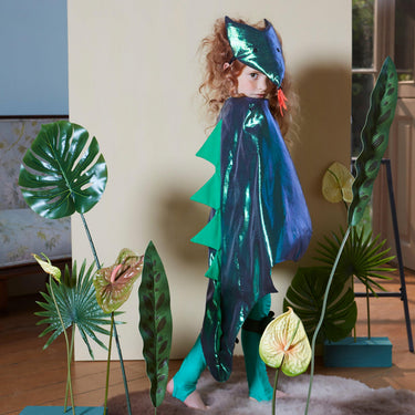 Child in a blue and green dinosaur costume standing among artificial plants indoors.