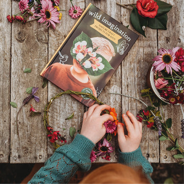 A book called Wild Imagination on a wooden table with flowers and leaves strewn about and a child's hand making a flower crown
