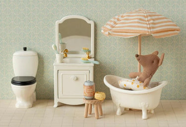A miniature dollhouse bathroom scene featuring a white clawfoot bathtub with a brown plush deer sitting inside under a striped peach and white umbrella, a white vanity cabinet with a mirror, and a white toilet against a green patterned wallpaper background.
