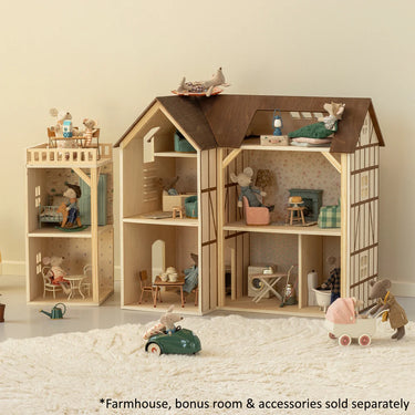 A large wooden farmhouse dollhouse with an attached bonus room and miniature furniture, toys, and accessories arranged inside and around it on a white textured rug. The scene is photographed in a clean indoor setting with a light wall background.
