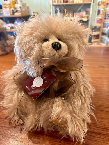 A fluffy beige stuffed teddy bear with shaggy fur, a black embroidered nose, and a sheer brown ribbon bow tied around its neck, with a product tag attached. The bear is sitting on a wooden table with a blurred store background and the letter B on its tag.