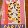 A greeting card with a colorful party design, wrapped in clear plastic and photographed on a wooden surface. The card features the text 'LET'S PARTY!' with smiling emoji-style faces wearing party hats.