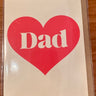 A square greeting card with a large red heart and the word 'Dad' in white, photographed on a wooden surface inside a clear protective sleeve.