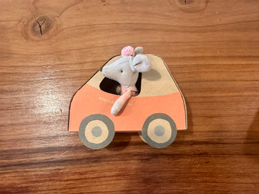 A maileg driver mouse wearing a pink headband,leaning out of her cardboard box shaped like a car, resting on a wooden tabletop.