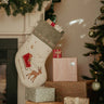 A Christmas stocking with a beige body, gray velvet trim, and an embroidered reindeer pulling a sleigh hangs on a fireplace mantel beside wrapped gifts and a decorated Christmas tree.