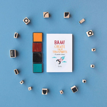 A flat-lay of a white art print with the text 'BAM! CREATE YOUR TRANSPORTS' and a small illustration, surrounded by small wooden stamp blocks arranged in a circle on a blue background. To the left of the print are four vertical color blocks in orange, rust red, black, and teal.
