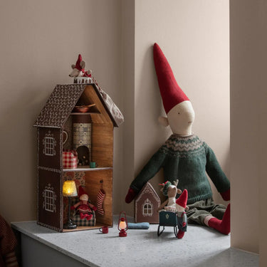 Gnome figurine with a red hat and green sweater next to a gingerbread house with small figures on a neutral background.
