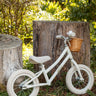 A white balance bike with a small wicker basket on the front, displayed outdoors against a weathered tree stump in a garden setting.