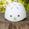 A white safety helmet with ventilation holes and a green circular 'Z' logo on the front, resting on a rustic wooden log with a blurred green forest background.