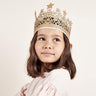 A young girl with brown hair wearing a gold glitter crown with star cutouts, looking to the side against a plain off-white background.