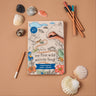 A book with a child's hand surrounded by shells and pencils on a pink background