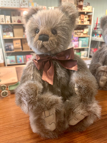 A plush teddy bear with shaggy gray-brown fur, a dark brown bow tie, and cream-colored stitched paws, sitting on a wooden table in a shop setting.