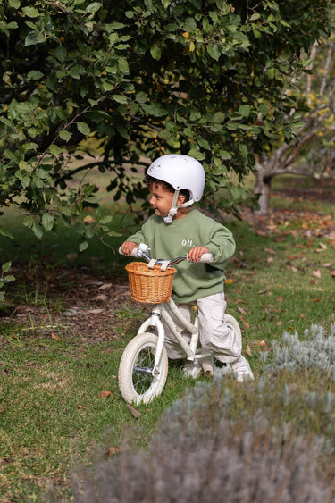 A toddler wearing a white helmet and a sage green sweatshirt rides a white balance bike with a wicker basket on a grassy path under a tree.