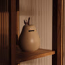 A Ferm Living pear-shaped ceramic coin purse in a warm beige color, styled on a wooden shelf against a ribbed beige background.