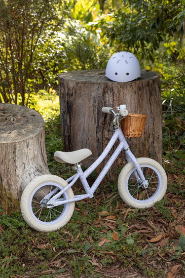 A small children's balance bike in a pale lavender-purple color with cream tires, a cream saddle, and a small wicker basket on the front, displayed outdoors on a grassy ground between two tree stumps with a matching helmet on top.