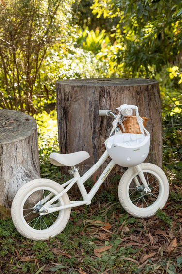 A small white balance bike with a wicker basket on the front, displayed outdoors on grass in front of tree stumps with a lush green garden background.