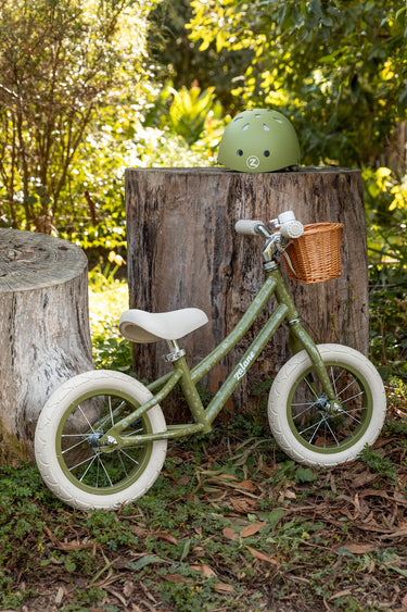 An olive green balance bike with white tires and a small wicker basket on the handlebars, parked outdoors against a tree stump with a matching green helmet resting on top. 
