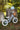 A small balance bike with a light blue frame and cream-colored tires, displayed outdoors on a grassy ground between two tree stumps. A matching light blue helmet and a small wicker basket sit on the stumps.