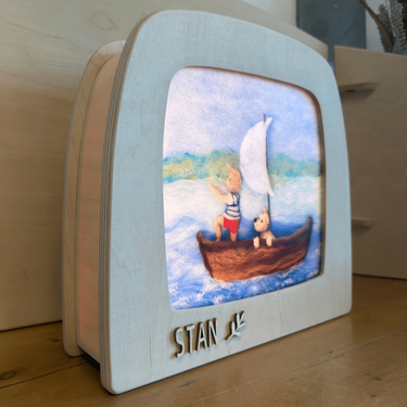 A personalized wooden keepsake box with a light-colored frame and a clear window showing a painted scene of a child in a sailboat with a teddy bear. The name 'STAN' is cut into the front, and the box is resting on a wooden surface.