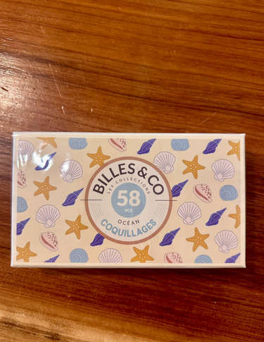 A rectangular box of Billes & Co. Ocean Coquillages (58 pieces) featuring a beige background decorated with colorful illustrations of starfish, seashells, and waves, placed on a wooden table.